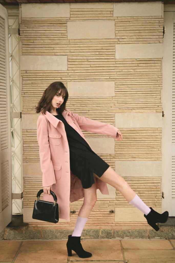 Fashion jeans model in pink coat and black dress striking a dynamic pose indoors with stylish black heels.